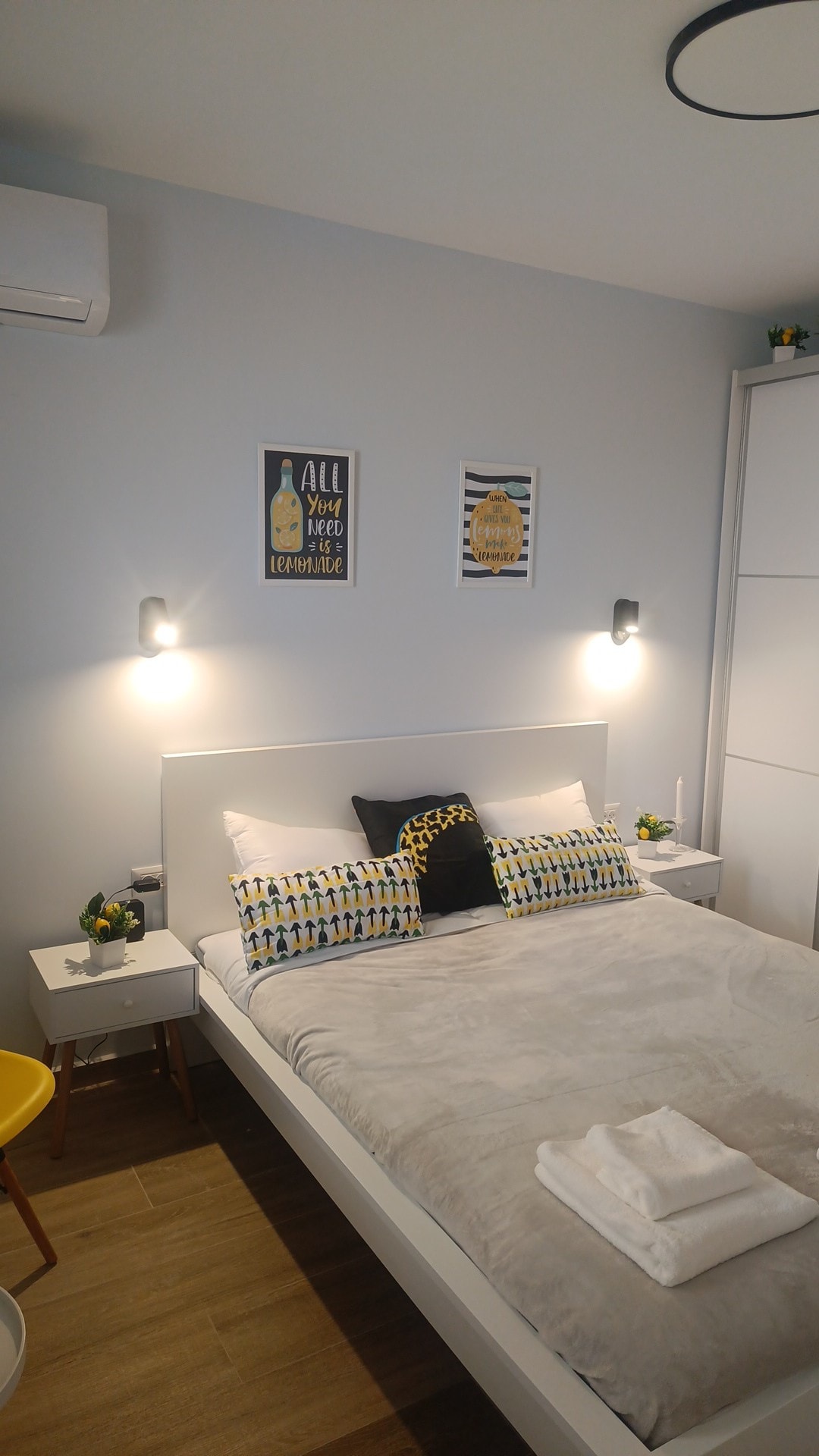 A well-lit bedroom features a spacious bed covered with a light gray blanket. Decorative pillows with various patterns are arranged against the headboard. Modern wall sconces illuminate the space. Artwork is displayed on the walls, complemented by small potted plants on a nearby table.