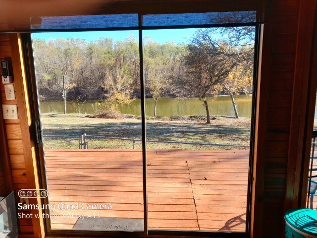 A large glass sliding door reveals a serene view of the Canadian River and surrounding trees. A wooden deck extends outward, providing direct access to the natural landscape. The calm waters reflect various shades of green from the foliage.