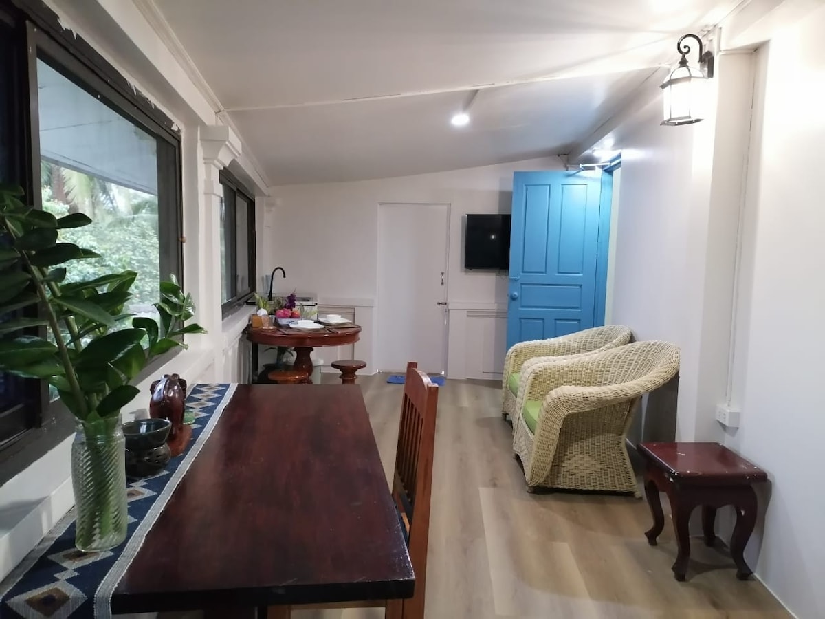 A well-lit room is furnished with a dining table and chairs, accompanied by two comfortable armchairs. A small side table sits beside the chairs. A vibrant plant is positioned on the table, with natural light filtering through the large windows.