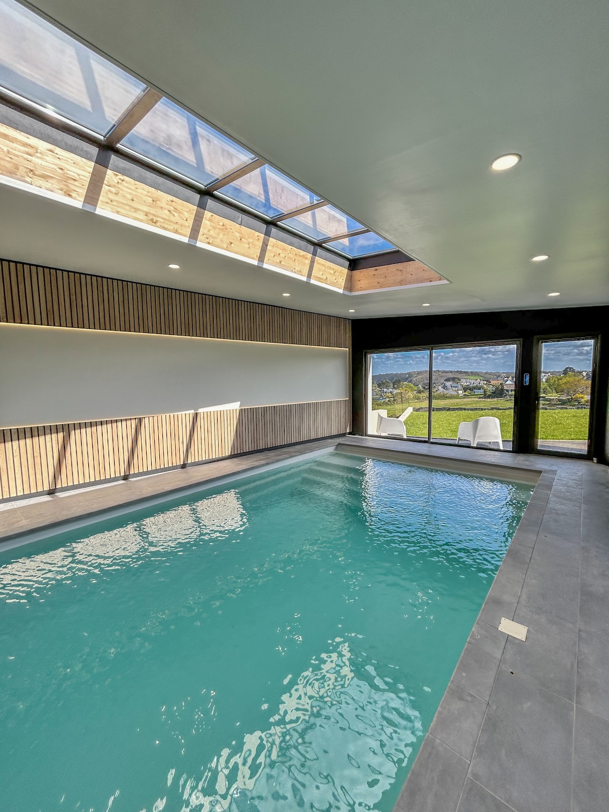 An indoor heated pool occupies the center of the spacious area, with large glass doors offering views of the sea. A skylight allows natural light to fill the room, creating a bright and inviting environment.