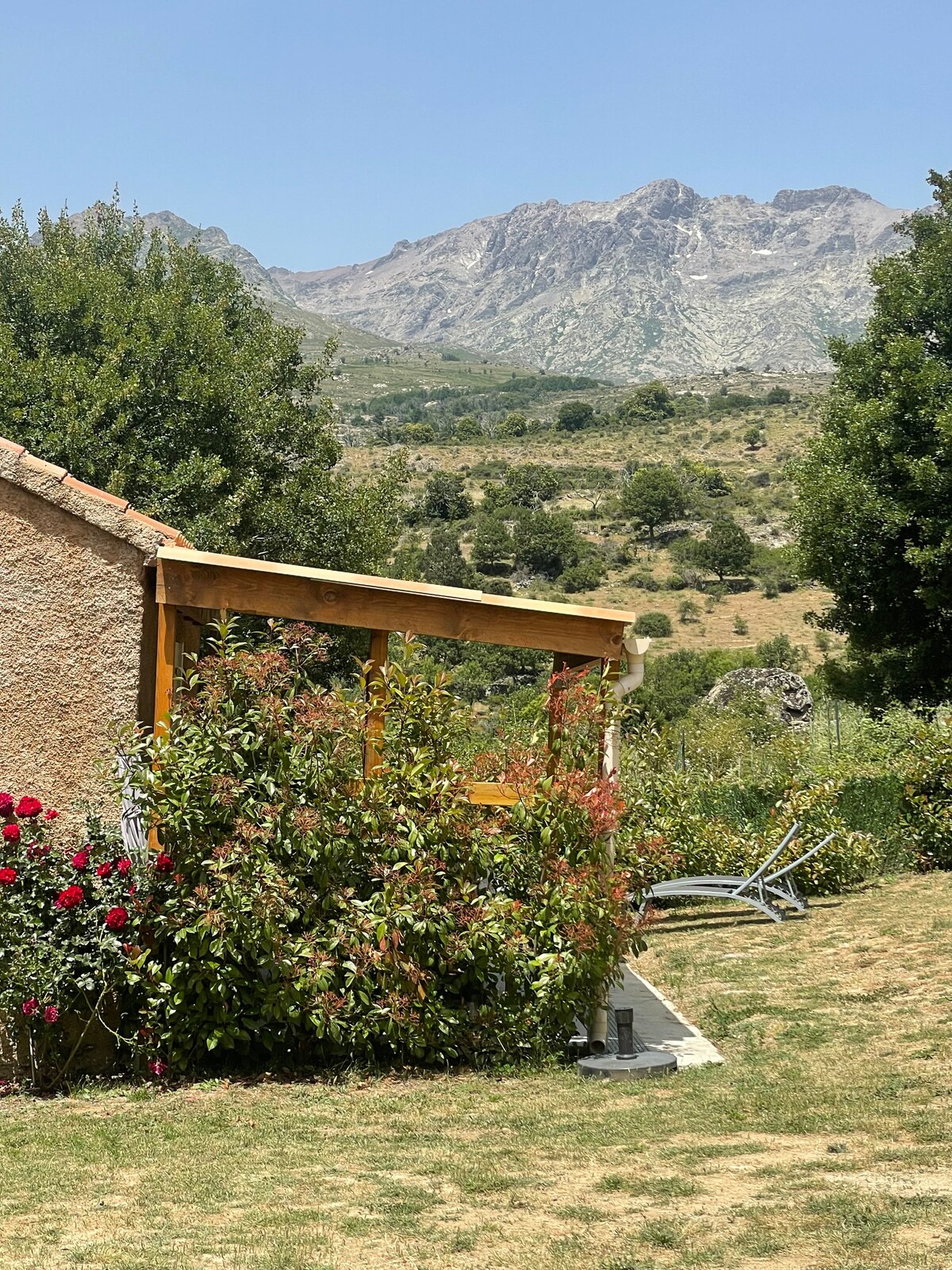 A rustic exterior view of a house is presented, surrounded by lush greenery and blooming flowers. The backdrop features majestic mountains, highlighting the natural landscape. Sunlight bathes the area in warmth, and a shaded patio area is visible, offering a place to relax outdoors.