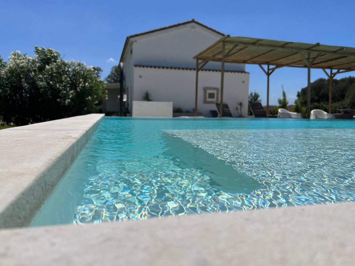 The image displays a clear, inviting swimming pool with shimmering water reflecting the sky. A spacious villa is positioned in the background, complemented by a shaded outdoor seating area under a pergola. Lush greenery frames the poolside, enhancing the serene environment.