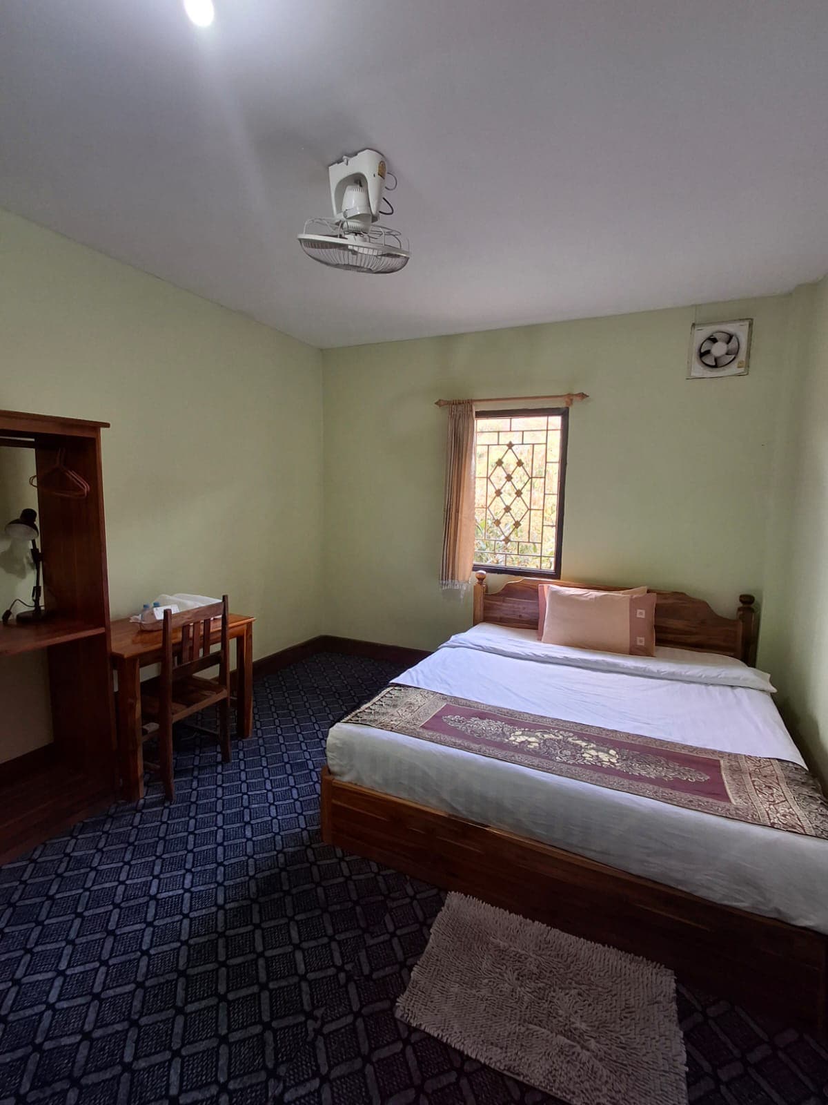 The room features a spacious bed with a decorative blanket. A small desk and chair are positioned near a window that allows natural light. The walls are painted in a soft green shade, and a ceiling fan is mounted for airflow. A closet stands to one side.