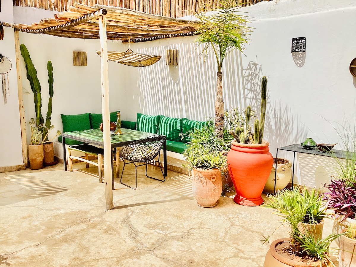 A serene outdoor seating area features a wooden pergola providing shade over a green bench and a low table. Decorative plants are arranged in terracotta pots, creating a natural ambiance. Textured walls complement the space, enhancing a relaxing atmosphere for guests.
