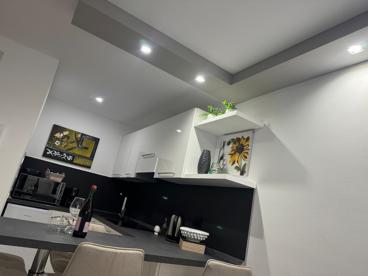 The kitchen area features modern white cabinetry against a sleek black backsplash, complemented by ambient lighting. A dining table is positioned nearby, with stylish chairs and a bottle of wine placed on the surface, inviting a cozy meal. Decorative elements include a potted plant and wall art.