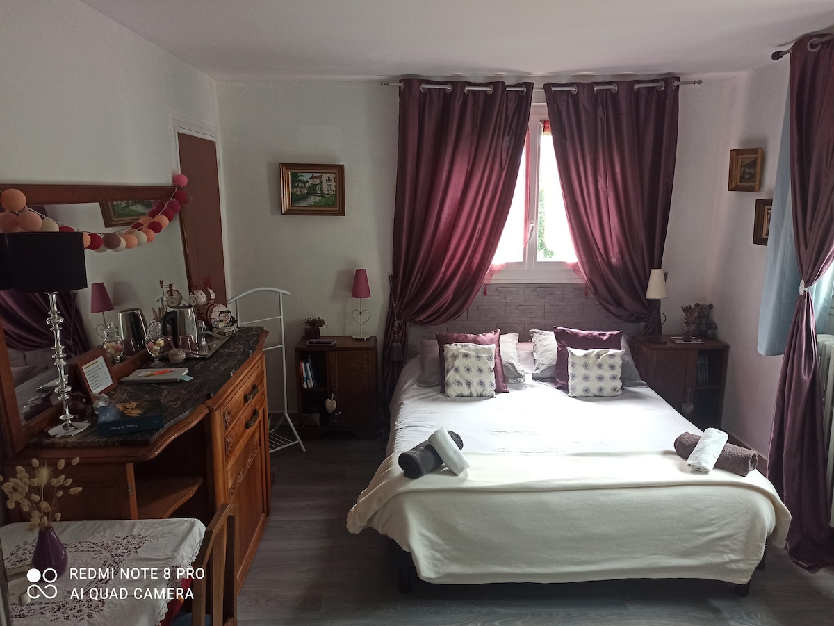 A cozy bedroom is presented with a spacious bed dressed in neutral linens. Dark curtains frame a bright window, contributing to the calm atmosphere. A wooden dresser and bedside tables flank the bed, while decorative items and small lamps add warmth to the space.