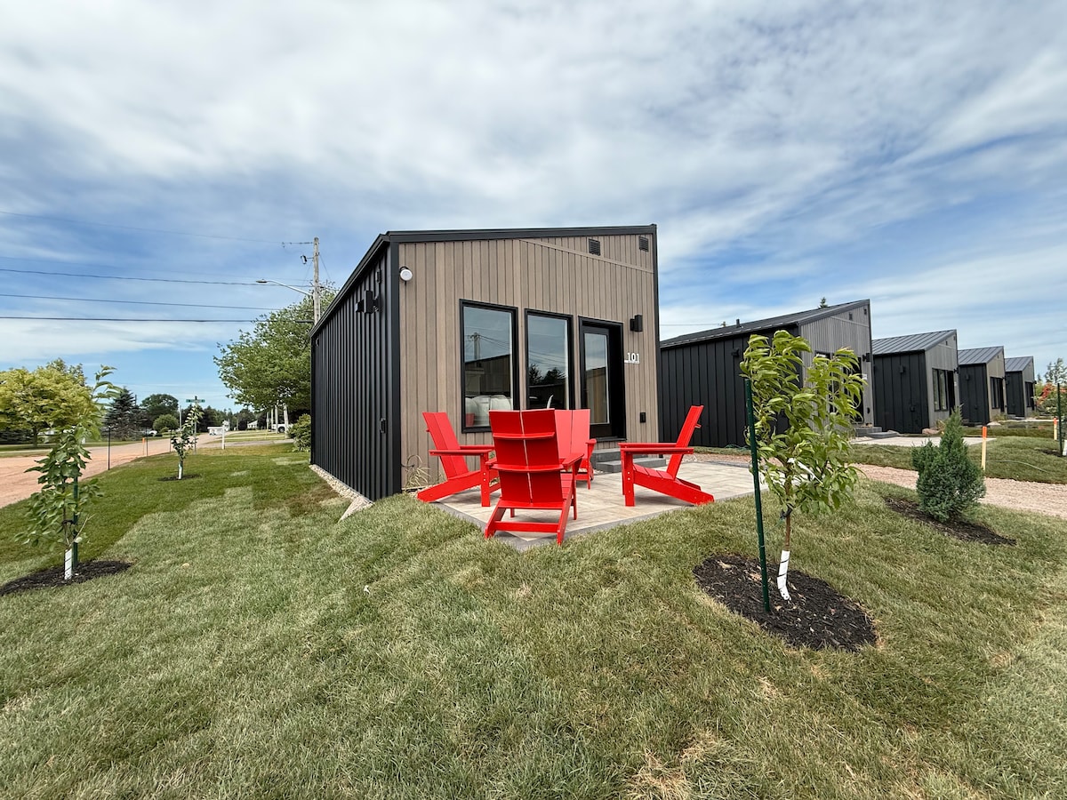 A modern cottage is depicted with large windows and a patio area. Red outdoor chairs are arranged around a circular table on a concrete patio, surrounded by neatly maintained grass and small trees. Additional cottages are visible in the background.