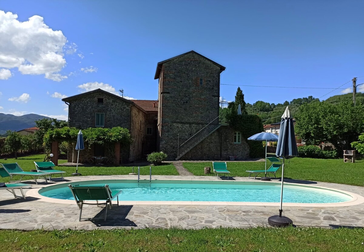The stone house features a medieval tower and is surrounded by a spacious garden. A private swimming pool is framed by sun loungers, offering a relaxing space to enjoy the view of green hills. The clear sky adds a bright ambiance to the setting.