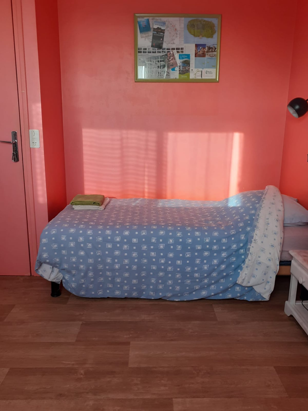 A single bed is positioned against a vibrant red wall, complemented by a light blue patterned blanket. A small bedside table holds a lamp, and a green towel is neatly placed on the bed. Natural light creates a pattern on the wall through the window.