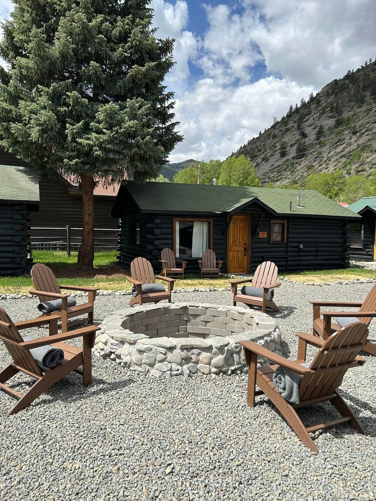 A communal fire pit is surrounded by six wooden Adirondack chairs, inviting relaxation. A natural stone border encircles the fire area, set on a gravel ground. Lush green trees and a mountainous backdrop complete the scene, highlighting the rustic charm of the log cabin surroundings.