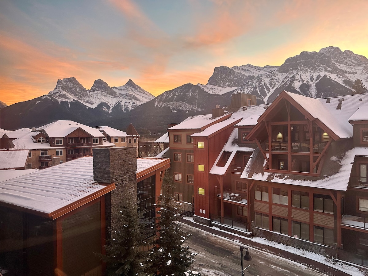 The view from the suite showcases the majestic Three Sisters mountain range at sunrise, framed by a serene winter landscape. Snow covers the rooftops of nearby buildings, with trees lining the street beneath. Soft hues of orange, pink, and blue populate the sky.
