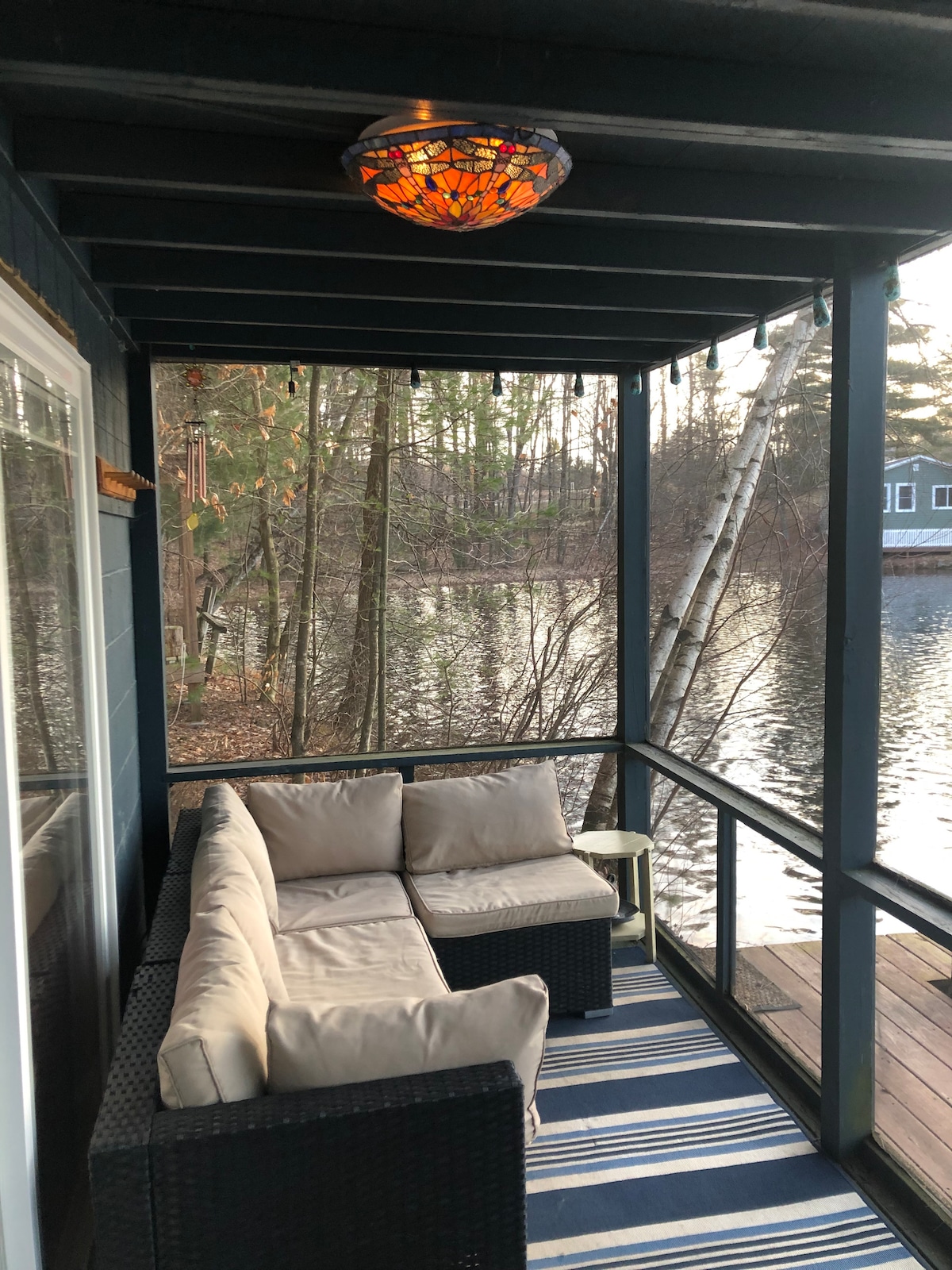 A screened-in patio area features a dark wicker seating arrangement with light-colored cushions. A small side table is positioned nearby, and elegant lighting hangs from the wooden ceiling. The serene water view reflects the surrounding trees, enhancing the peaceful ambiance.