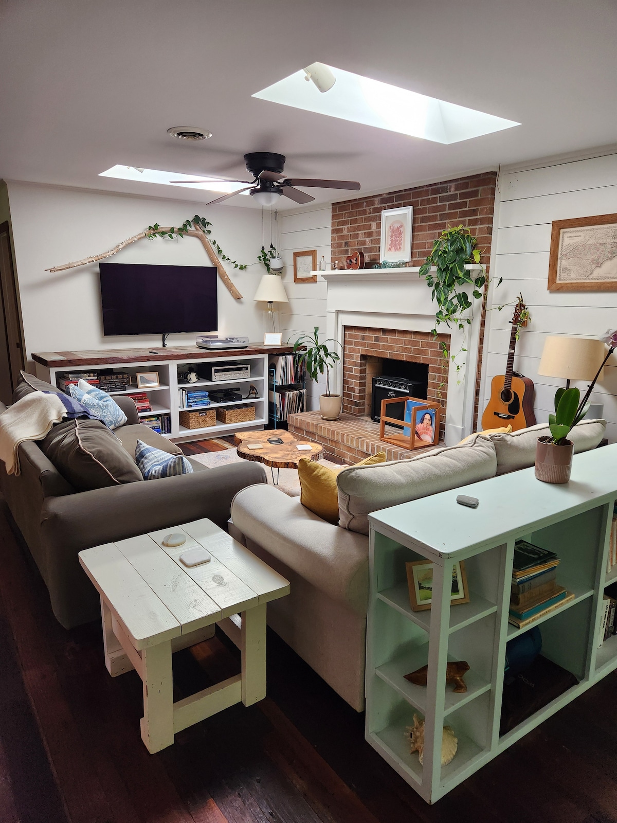 A cozy living room features two comfortable sofas positioned around a central coffee table. A television is mounted above a brick fireplace, while a guitar leans against the wall. Natural light enters through a skylight, and decor includes plants and framed art.
