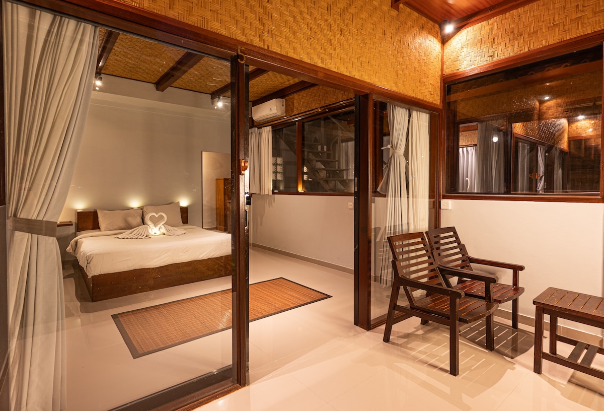 The bedroom area features a spacious bed adorned with decorative towels shaped into a heart. Natural light filters through large windows covered with soft curtains, while wooden accents contribute to a warm ambiance. A small seating area with two chairs is positioned nearby.