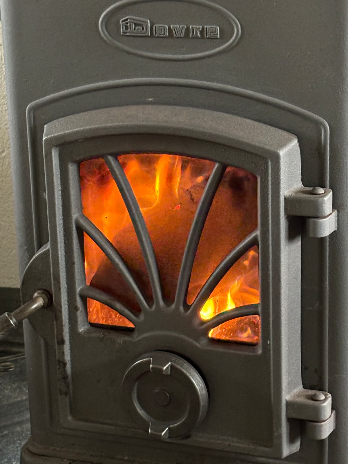 A wood-fired stove is depicted, featuring a decorative front with an arched design. Flames are visible through the glass door, providing warmth and comfort to the cabin's interior. The stove is designed for efficient heating, enhancing the cozy atmosphere of the rustic space.
