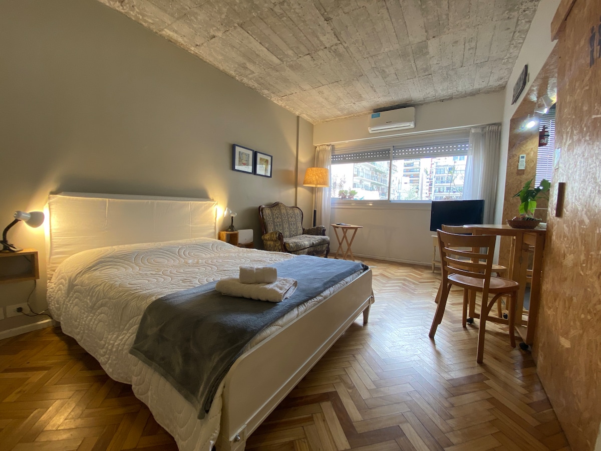 A spacious sleeping area is showcased, featuring a large bed with a light-colored quilt and soft pillows. A comfortable armchair is positioned beside a small table, with natural light filtering through a large window adorned with sheer curtains. An air conditioning unit is mounted above.