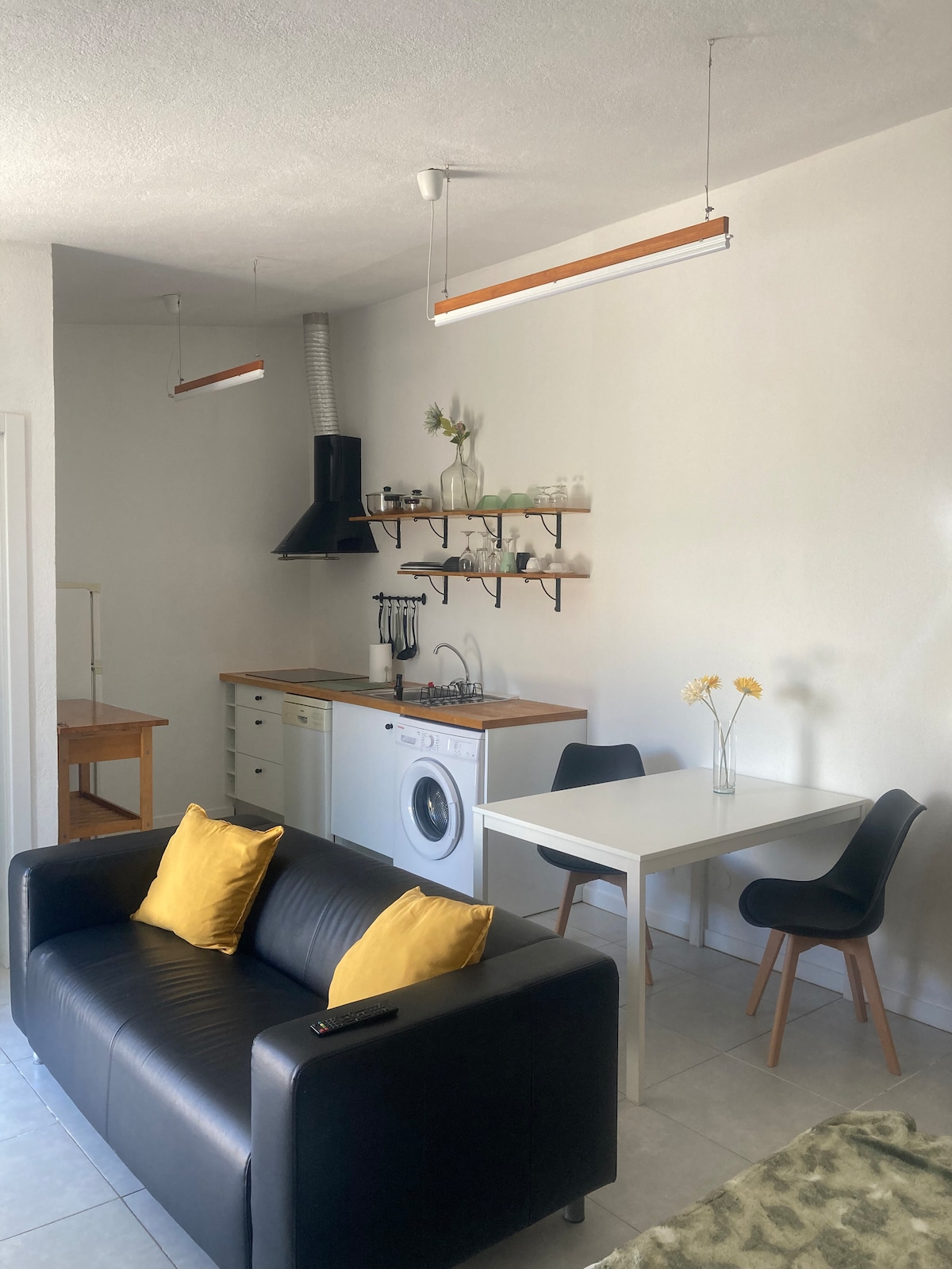 A modern open-plan kitchen and living area are presented, featuring a sleek black sofa with yellow cushions. A small dining table sits next to a washer, with shelving above displaying kitchen essentials. Light is provided by overhead fixtures, enhancing the organized space.