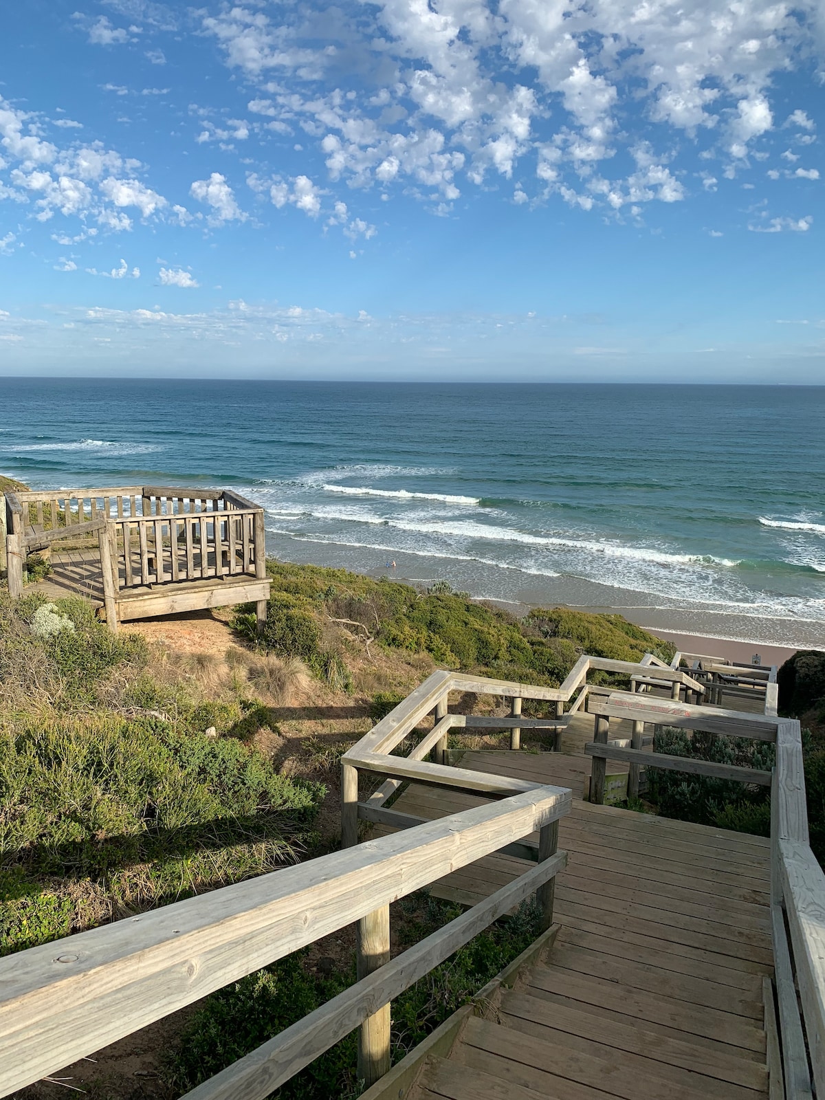 Wooden stairs wind down the hillside towards the ocean, flanked by greenery. Overlook platforms provide views of the waves crashing against the shore, under a clear blue sky with scattered clouds.