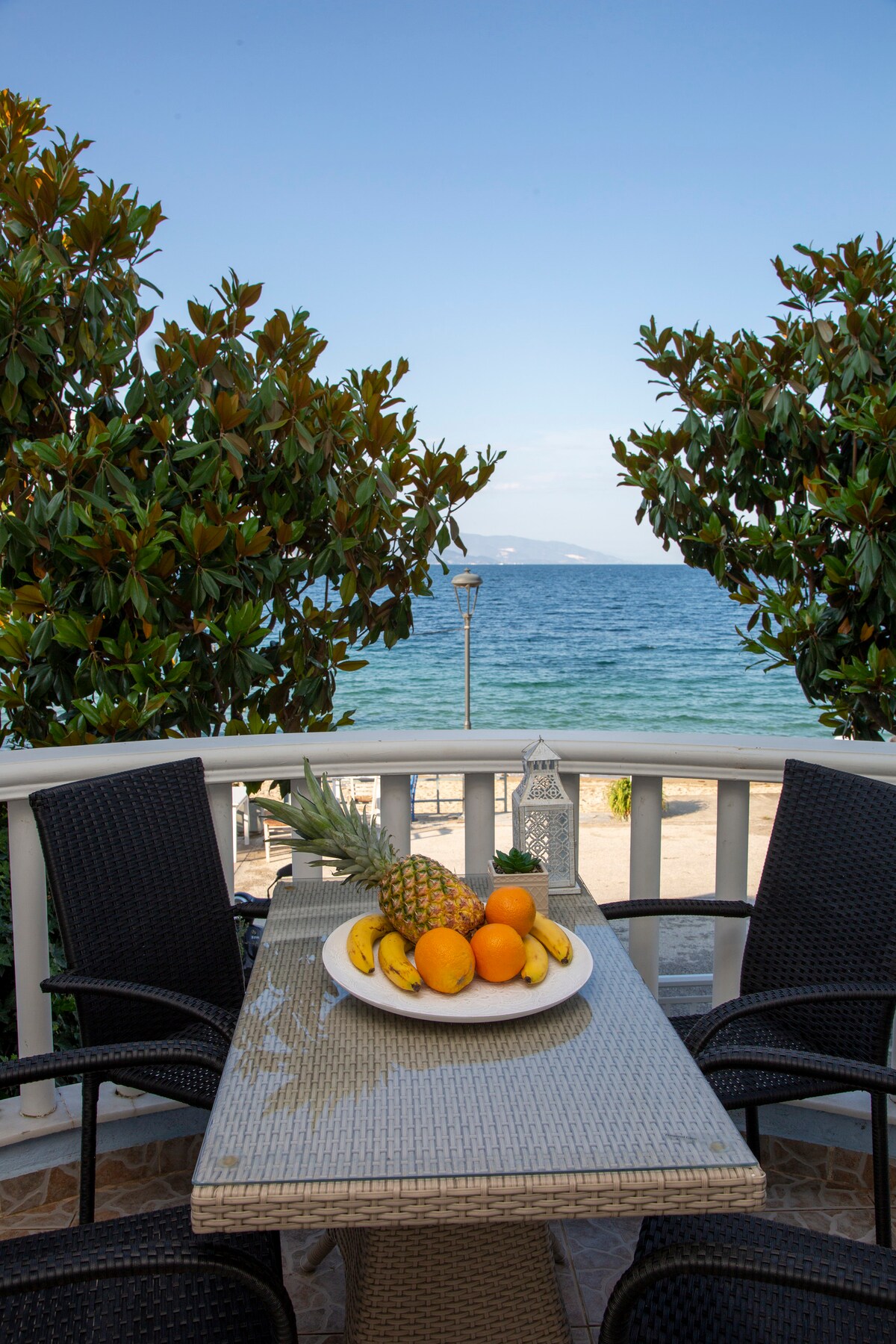 A dining table is set on a balcony, surrounded by dark wicker chairs. A bowl of fresh fruit, including oranges, bananas, and a pineapple, is placed in the center. Lush green trees frame the view of the calm sea and clear blue sky.