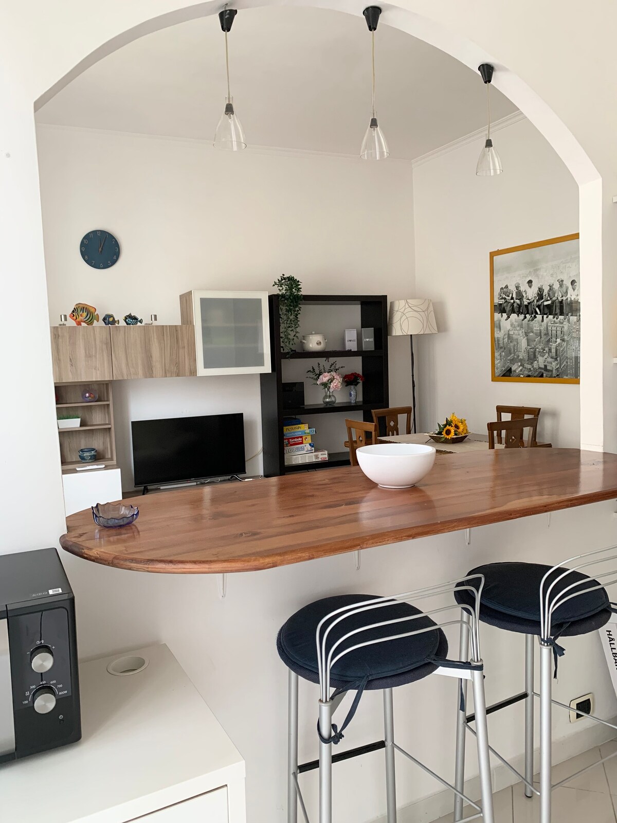 A modern dining area features a wooden countertop with three high stools. Behind, a wall-mounted TV is visible alongside a minimalist shelf holding decorative items. A framed artwork is positioned above the bar area, and a small clock hangs on the wall, contributing to the room's organized layout.