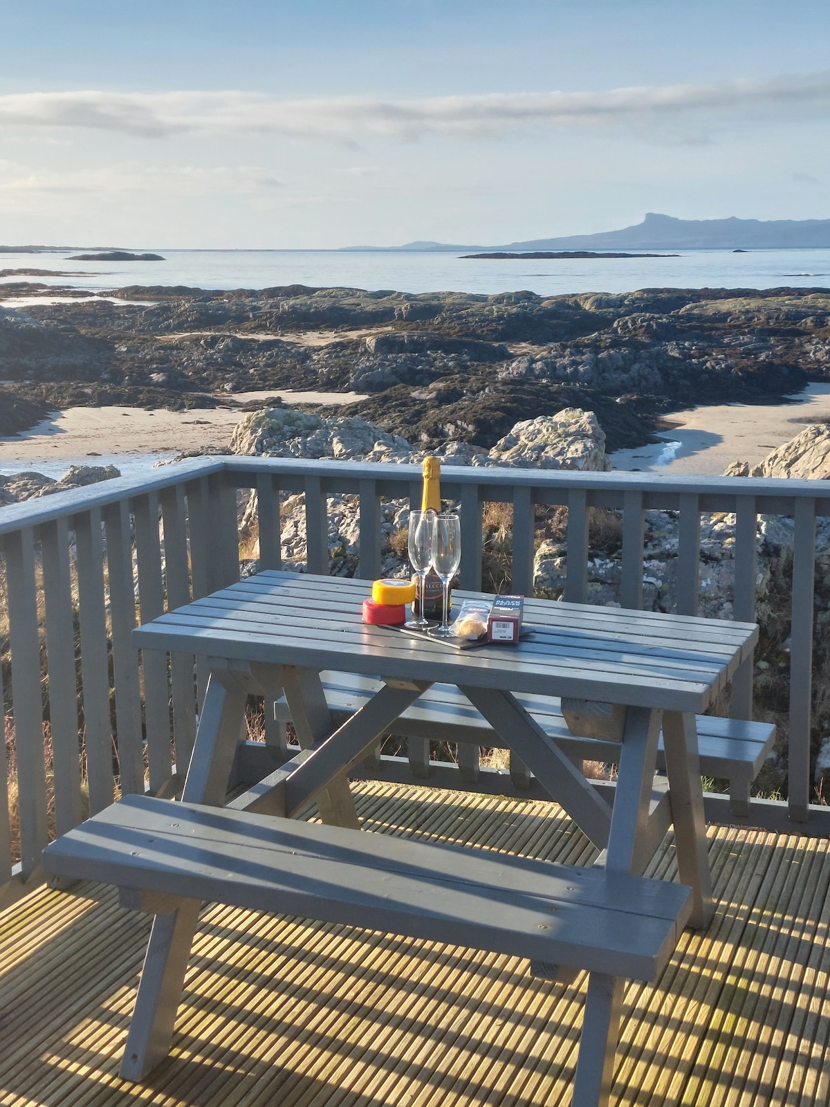 An outdoor picnic table made of wooden planks is positioned on an elevated decking area, offering views of rocky coastal landscapes and distant islands. A bottle of sparkling wine and two glasses rest on the table, accompanied by small containers and snacks.