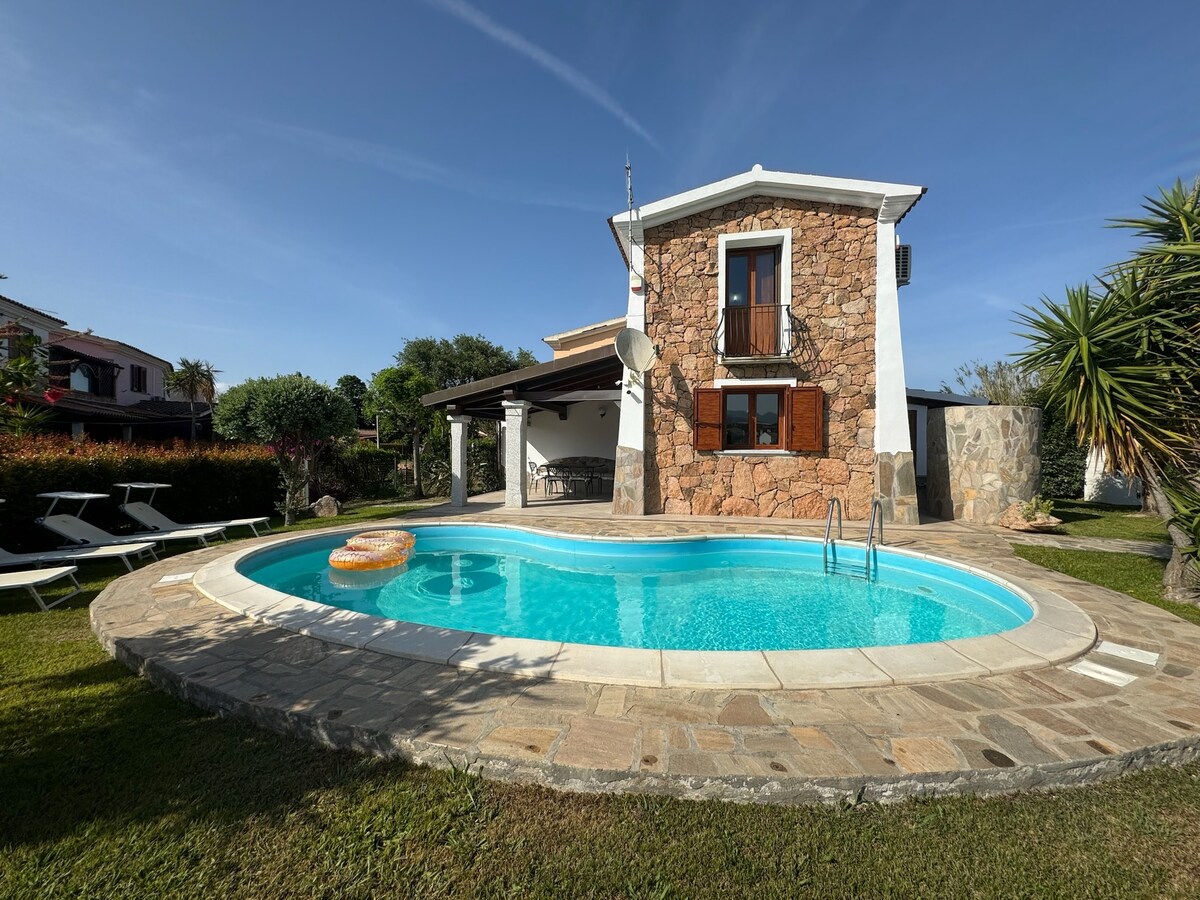 A charming villa features a rounded swimming pool surrounded by a stone patio. The building showcases a warm stone facade with wooden shutters. Lush greenery and sun loungers are positioned around the pool, creating an inviting outdoor space for relaxation.