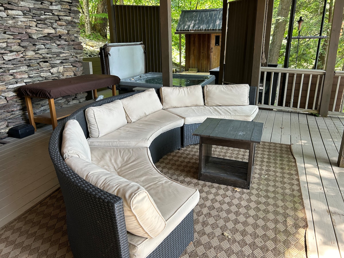 A spacious outdoor seating area features a curved sectional sofa with light fabric cushions, surrounded by natural stone walls. A wooden table sits in the center, and a hot tub is visible in the background, with greenery framing the space.