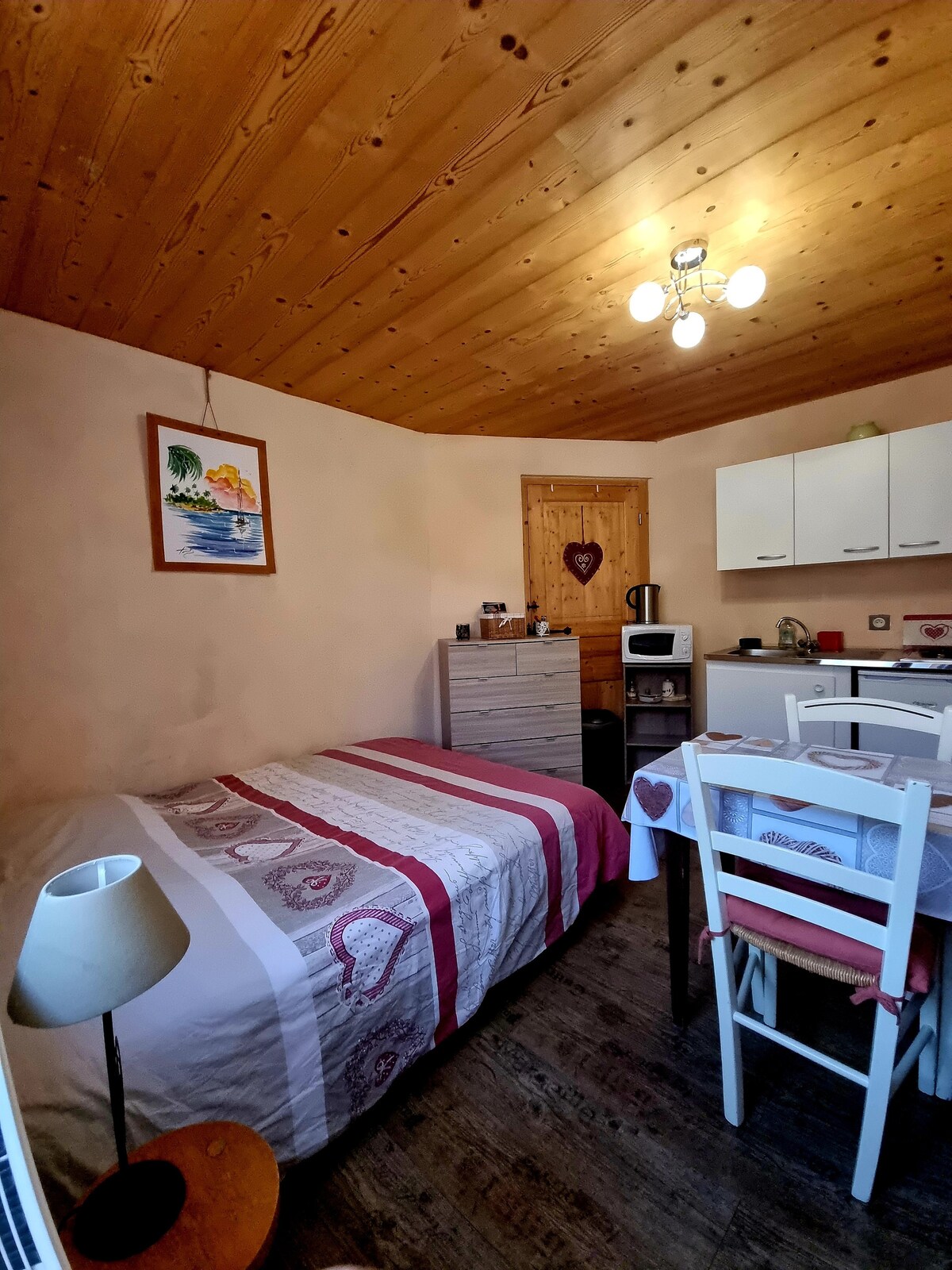 A cozy studio with wood-paneled walls and a ceiling adorned with soft lighting. A spacious bed is situated next to a drawer unit, with a kitchenette visible in the background. A small dining table and chairs are arranged near a window, fostering a comfortable atmosphere.