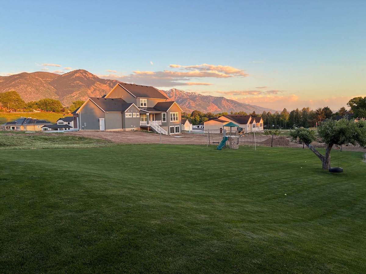 A spacious backyard is captured with a well-maintained lawn, leading to a modern gray house. The scenery features distant mountains under a beautiful sky, with soft clouds illuminated by the setting sun. Play equipment is visible on the lawn, enhancing the family-friendly atmosphere.