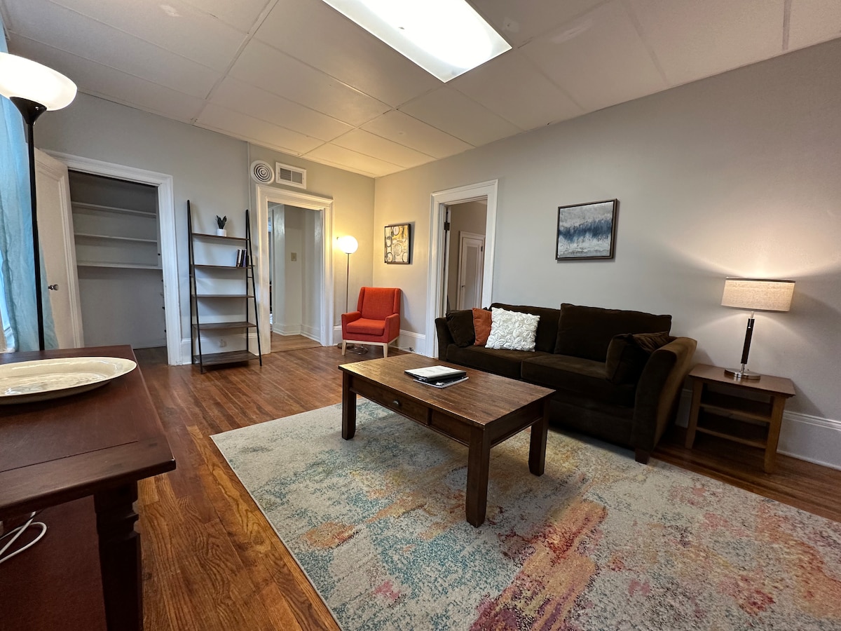 A cozy living area is showcased with a brown couch, an orange chair, and a wooden coffee table featuring a tablet. A rug with soft colors sits beneath, while floor lamps and a side table provide gentle lighting. Open doorways lead to additional spaces.