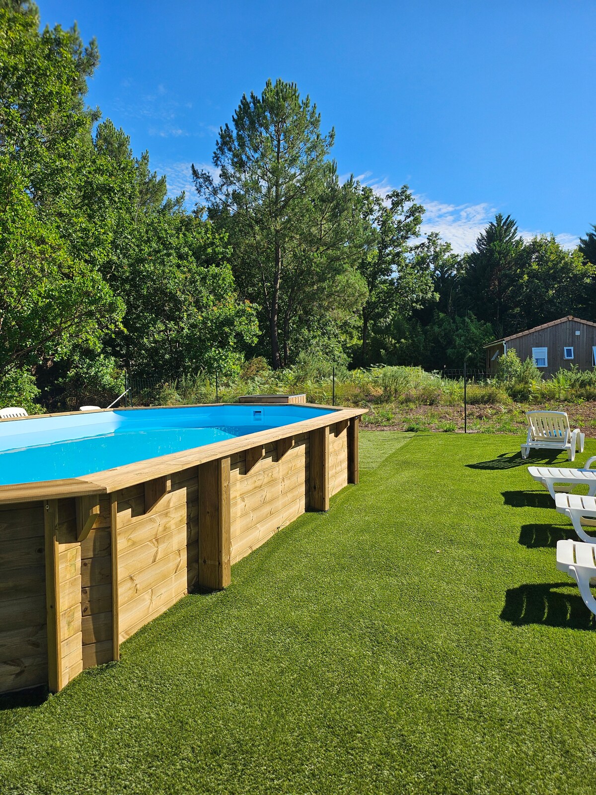An above-ground swimming pool with a wooden surround is set on a manicured green lawn. Several white lounge chairs are positioned nearby, offering a space to relax. Lush trees and greenery fill the background, providing a natural and serene setting.