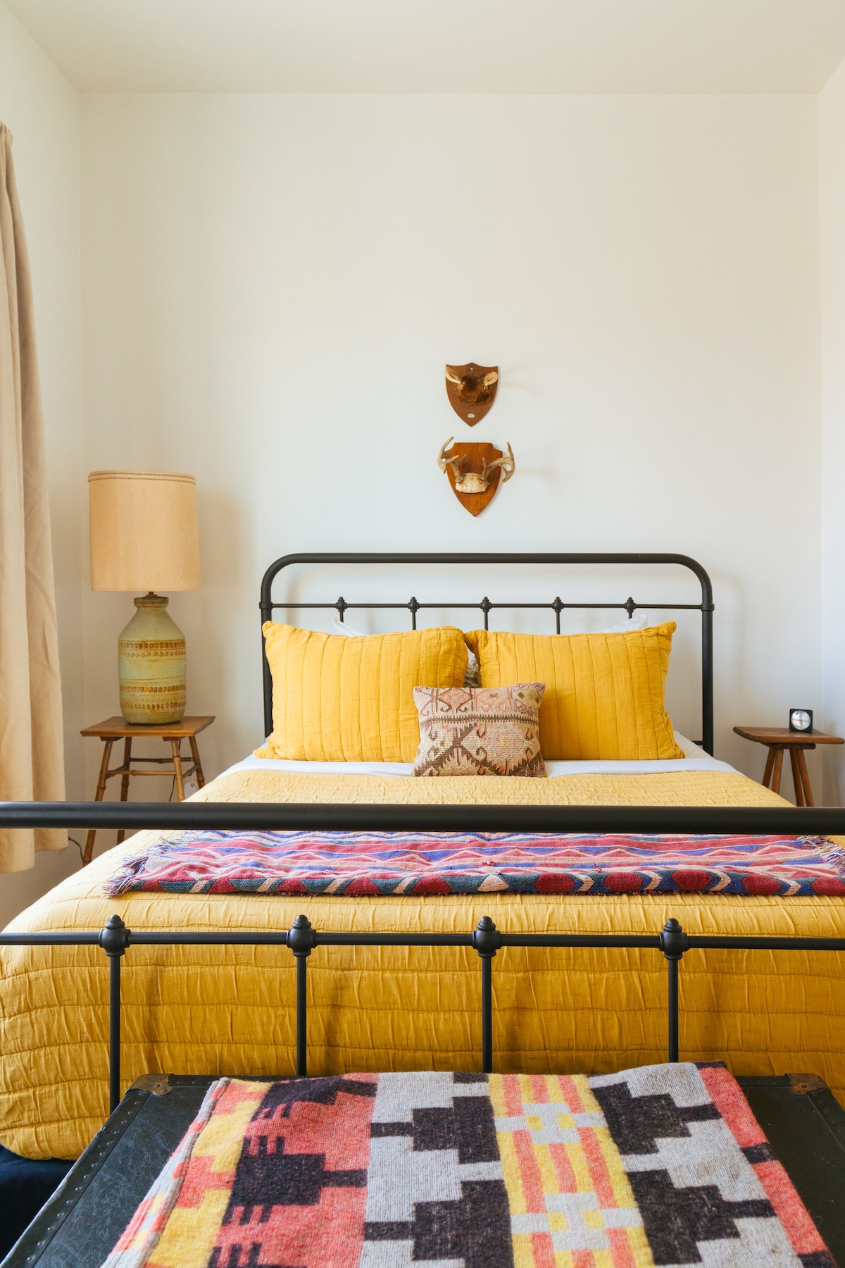 A cozy sleeping area features a black metal frame bed dressed in a vibrant yellow quilt. Decorative pillows and a colorful throw add texture. Nearby, a vintage lamp provides warm lighting, while two small wooden tables complete the inviting room decor.