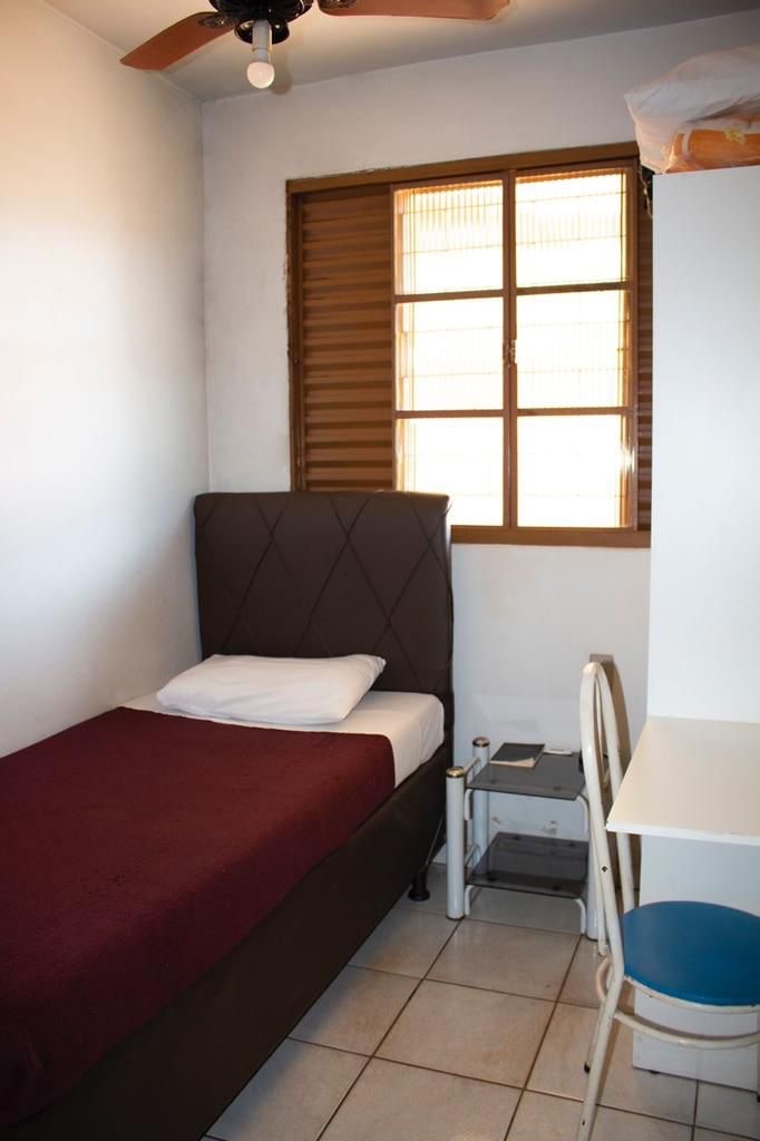 A compact bedroom features a single bed with a maroon blanket. Natural light enters through a wooden window with horizontal slats. A small nightstand is positioned next to the bed, complemented by a white desk and chair in the corner.