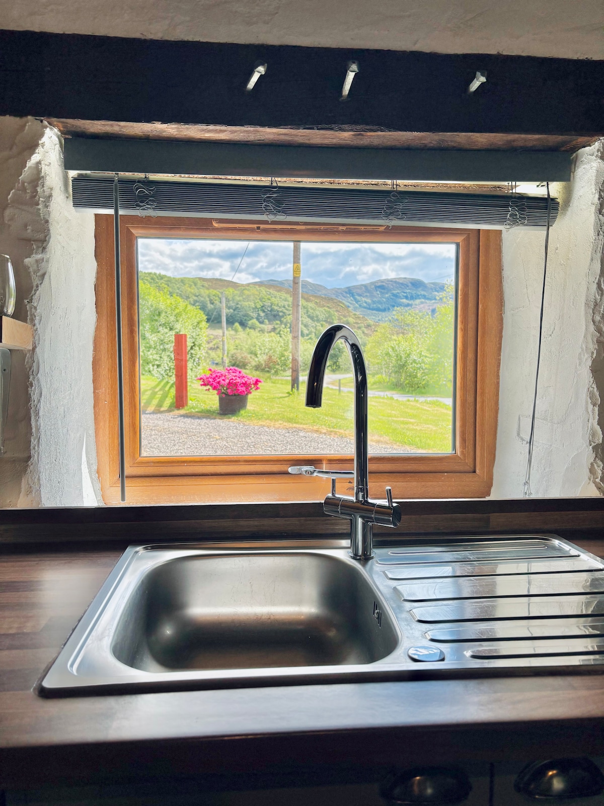 A modern kitchen sink is positioned in front of a window, providing a view of the outdoor landscape. Bright green hills and a flower pot with vibrant pink flowers are visible outside, contributing to a tranquil and inviting atmosphere.