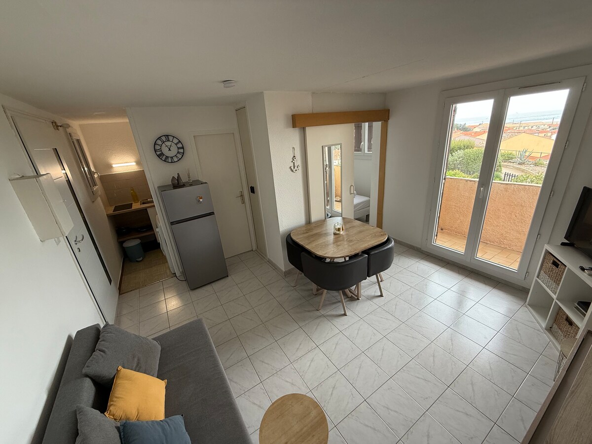 A bright and airy living space is visible, featuring a round dining table surrounded by four chairs. A comfortable gray sofa and a coffee table are positioned to one side. Large windows offer a view of the outdoors, enhancing the overall spaciousness.