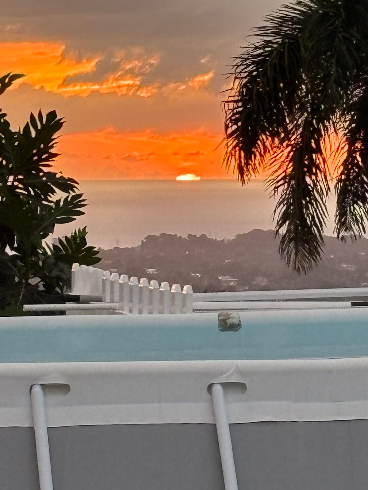 A scenic view of a vibrant sunset is captured over the horizon, with warm orange and yellow hues blending into the sky. The image features the edge of a swimming pool and tropical foliage, emphasizing the tranquil atmosphere of the location.