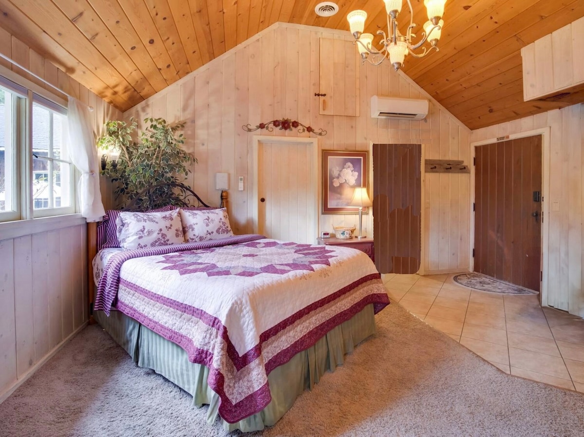 A cozy bedroom is characterized by a cathedral ceiling and features a quilted bed with neutral tones and floral accents. Natural light enters through the window, illuminating the warm, wooden interior. A small lamp is placed beside the bed, contributing to the welcoming ambiance.