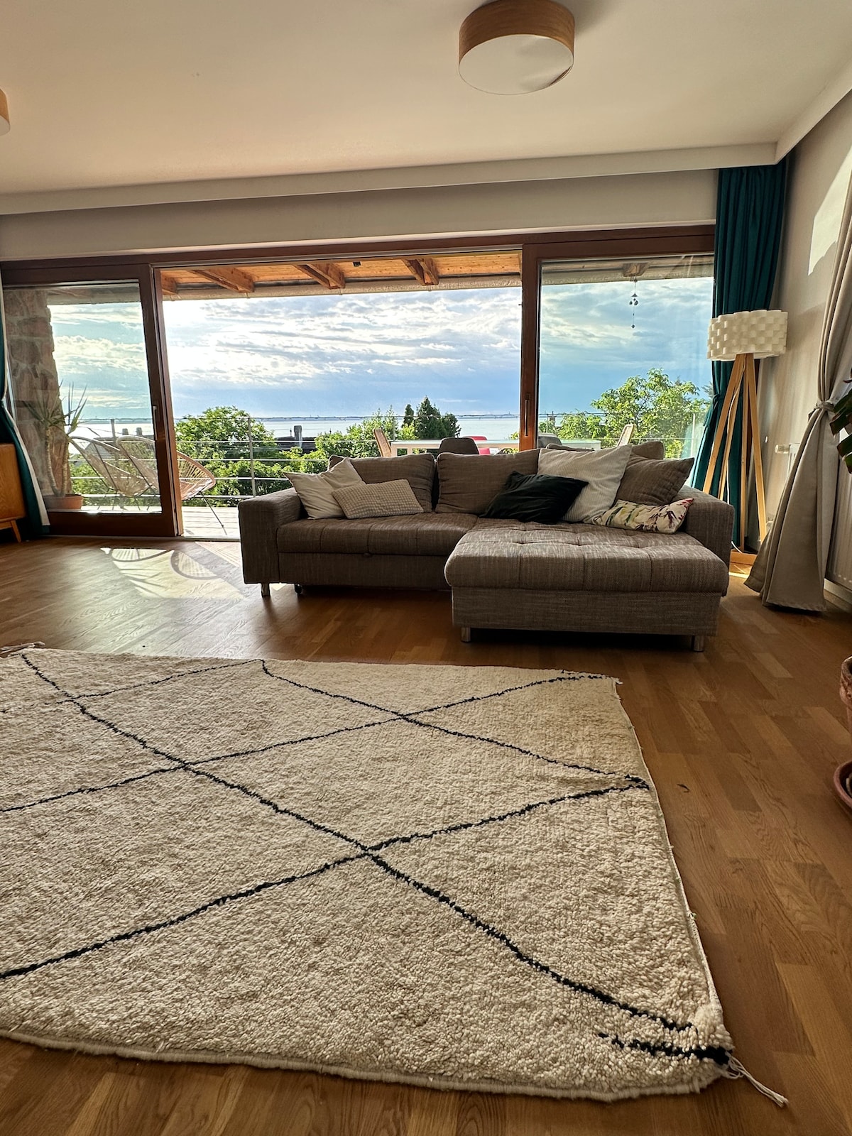 A spacious living area features large glass doors that open to scenic views. A comfortable couch with cushions is positioned beside a light-colored area rug. Natural light floods the space, enhancing the cozy atmosphere complemented by the surrounding greenery.