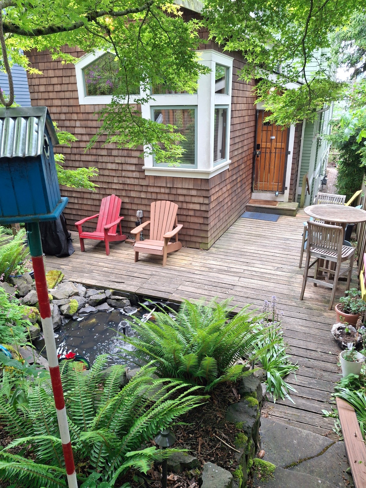 A private outdoor space features wooden decking surrounded by lush ferns and greenery. Two red adirondack chairs sit next to a small pond, creating a peaceful corner. A wooden table and chairs are positioned nearby, enhancing the inviting atmosphere of this secluded area.