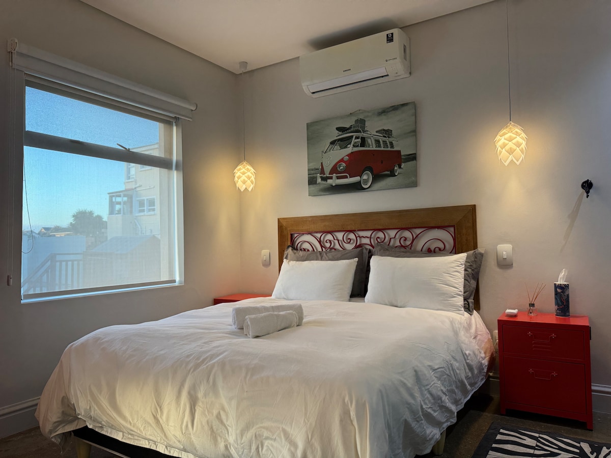 A well-lit sleeping area is showcased, featuring a spacious bed with luxury cotton bedding. A red nightstand is positioned beside the bed, and a colorful artwork depicting a vintage car adorns the wall above. Soft, diffused light filters through a window with sheer blinds.