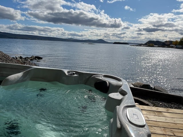 A hot tub is situated on a wooden deck overlooking the serene sea. Rippling water reflects sunlight, while distant hills rise against the cloudy sky. The scene emphasizes tranquility and invites relaxation.