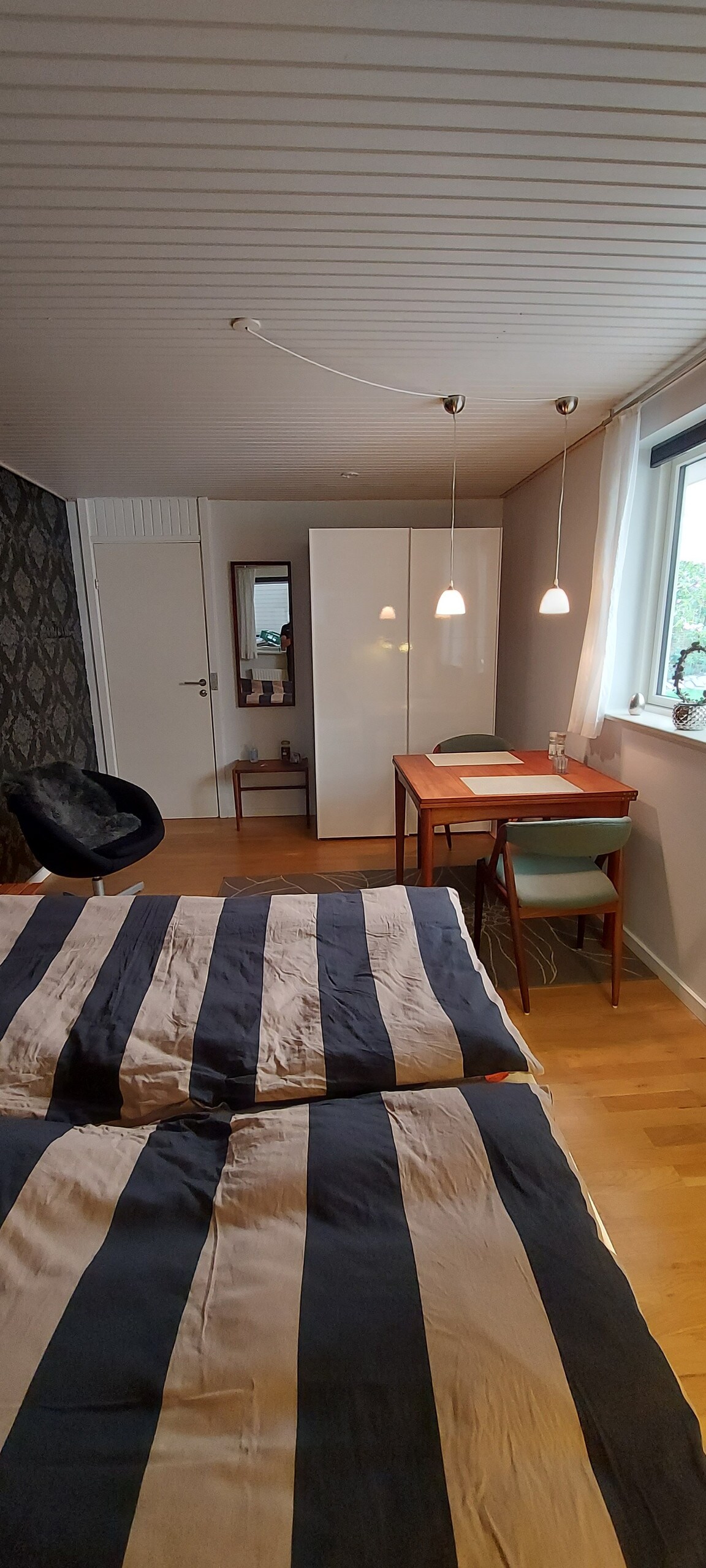 A cozy bedroom features a striped bedding set on a spacious bed. A small wooden desk with two chairs is positioned near the window, illuminated by soft light from pendant lamps. A full-length mirror and a wardrobe are also visible, along with a chair in the corner.