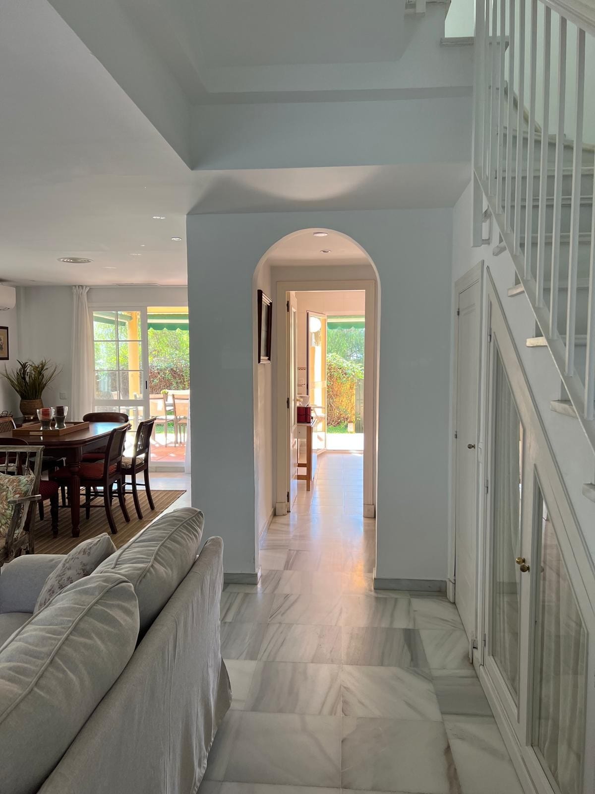 A light-filled corridor is seen, leading towards an open space. The walls are painted in soft white, enhancing the airy feel. A marble floor reflects the natural light, while an archway adds architectural interest. At the end, doors open to a patio, revealing greenery.
