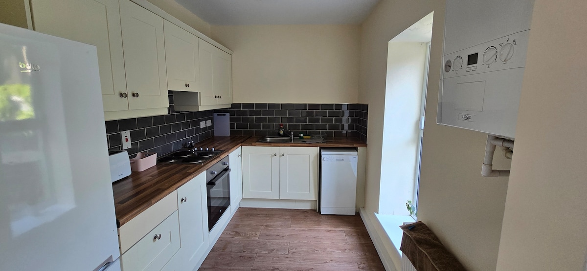 A modern kitchen features premium cabinetry in light tones and a wooden countertop. Stainless steel appliances, including an oven and a refrigerator, are visible. A window allows natural light into the space, and a washing machine is discreetly integrated into the cabinetry.