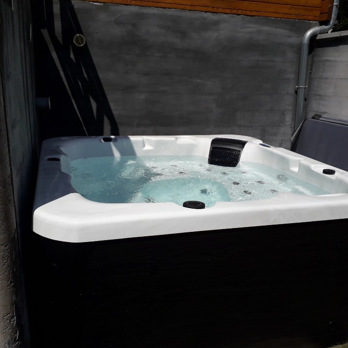 An outdoor jacuzzi is displayed, featuring a spacious design with a smooth white edge and a dark wooden panel base. Bubbles rise in the clear water, suggesting comfort and relaxation. The surrounding walls are simple and unadorned, offering privacy while enjoying the spa experience.