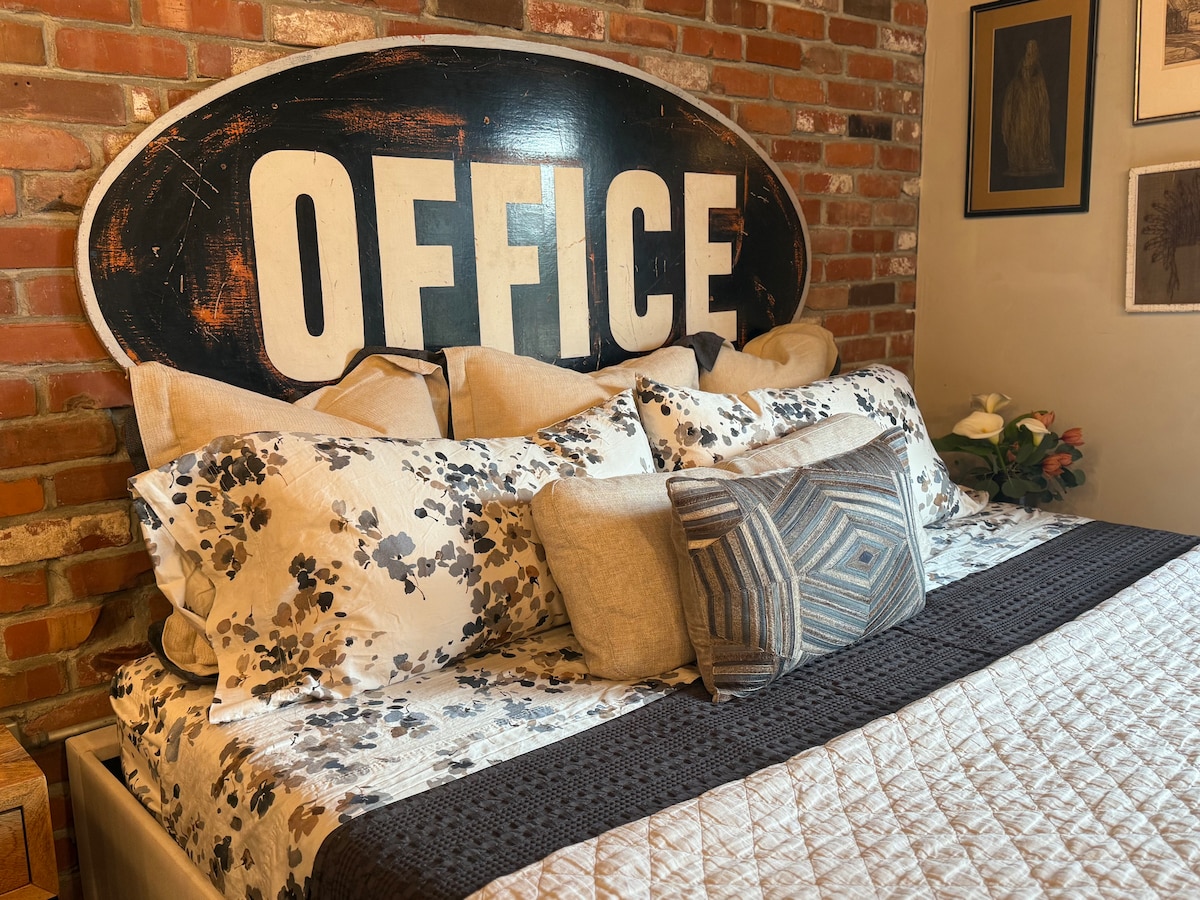 A cozy sleeping area features a comfortable bed adorned with a floral patterned duvet and a textured throw. The headboard, made from an old sign reading 'OFFICE,' adds a unique touch to the brick wall backdrop. Decorative pillows provide additional comfort.