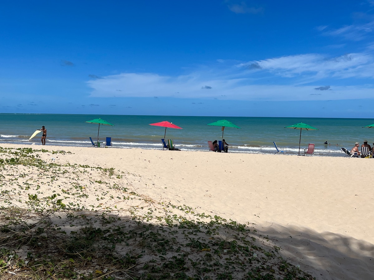 A sandy beach with gentle waves lapping at the shore is seen under a clear blue sky. Vibrant umbrellas in red and green offer shade to beach chairs positioned close to the water. Lush vegetation borders the beach area, creating a natural transition to the sand.