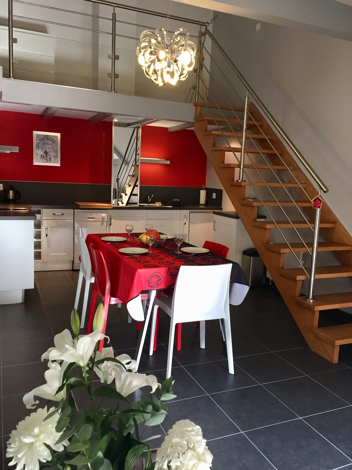 A dining area showcases a red table set for four, adorned with fruit and decorative items. Light is provided by a modern chandelier overhead, illuminating the sleek kitchen and staircase that lead to the upper level. Flowers are positioned in the foreground, adding a touch of nature.