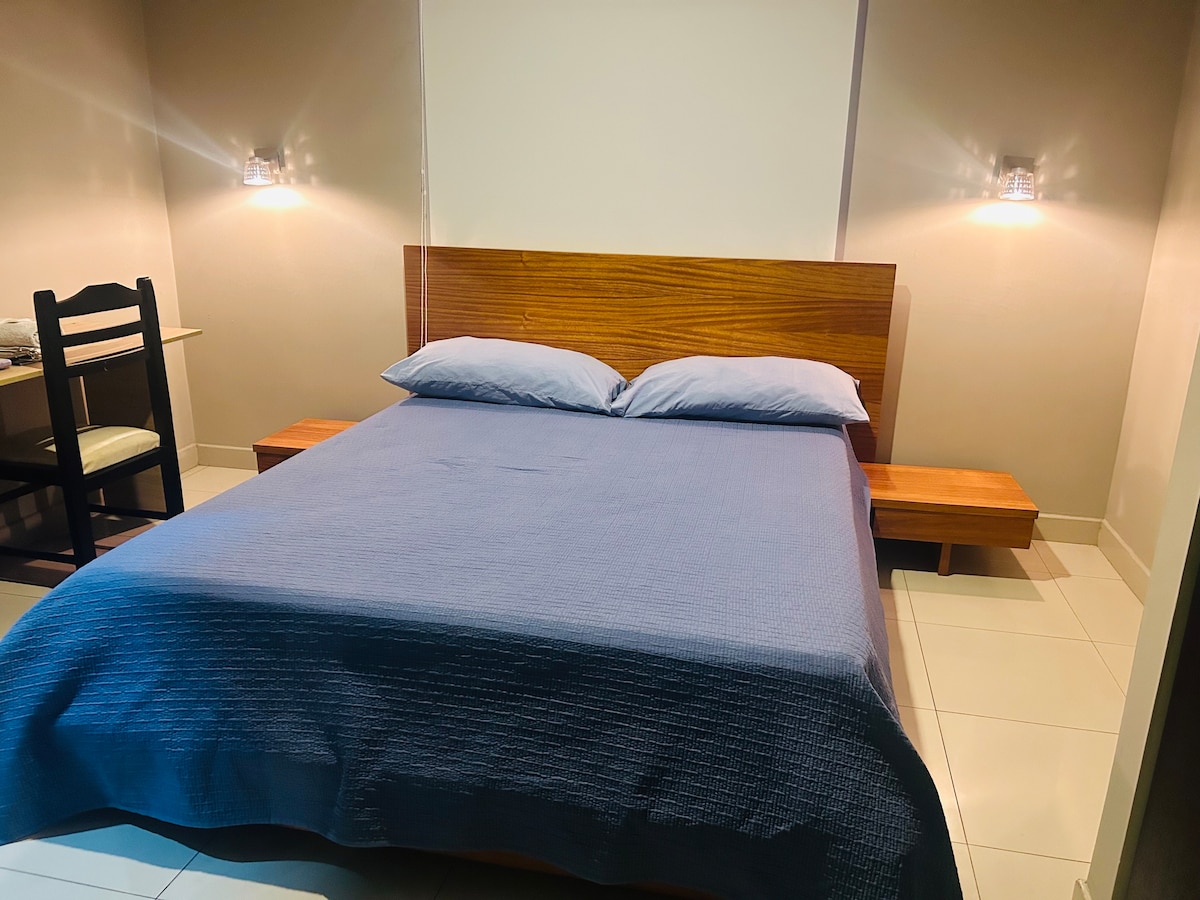 A spacious bed is centered against a smooth wall, complemented by bedside lighting. A minimalist wooden bench sits adjacent to the bed. A simple chair and small table are present in the corner, adding functionality to the room.