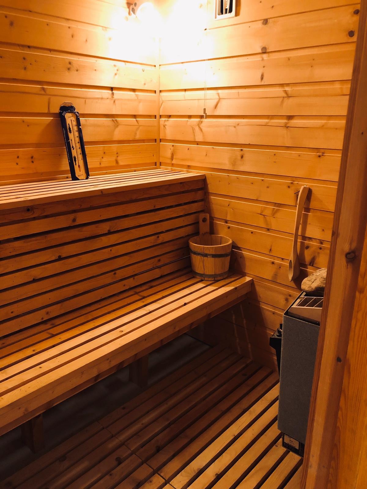 A warm sauna is lined with light wood paneling, featuring wooden benches arranged for seating. A small wooden bucket and a ladle are placed nearby, while a sauna heater rests at one side. Soft lighting creates a calming environment.